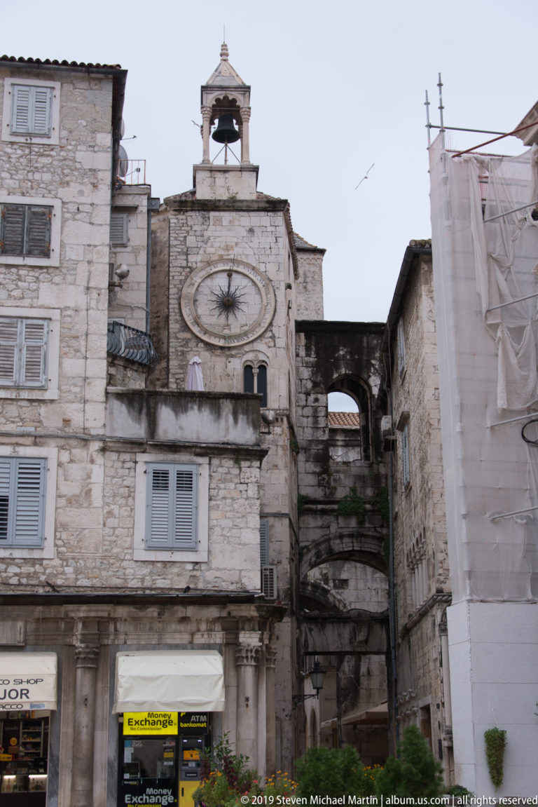 Weathered stone façade with circular opening, small rooftop bell cote, shuttered windows, shops, and adjoining historic masonry.
