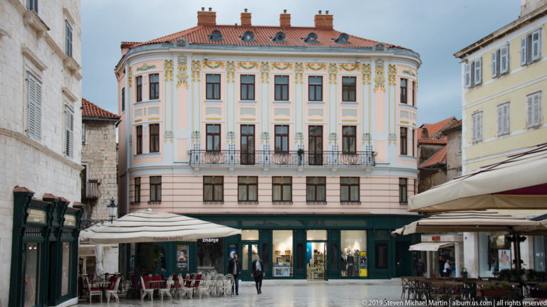 Symmetrical pale façade with painted floral ornament, balconies, shops, and a red tile roof.