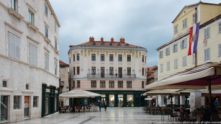 Decorated rounded corner building with balconies and shops facing a stone-paved pedestrian square.
