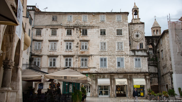 Historic stone buildings and café awnings on a Split square, with a clock tower rising at right.