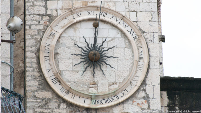 Large round clock with Roman numerals and a dark metal sunburst mounted on pale stone masonry.