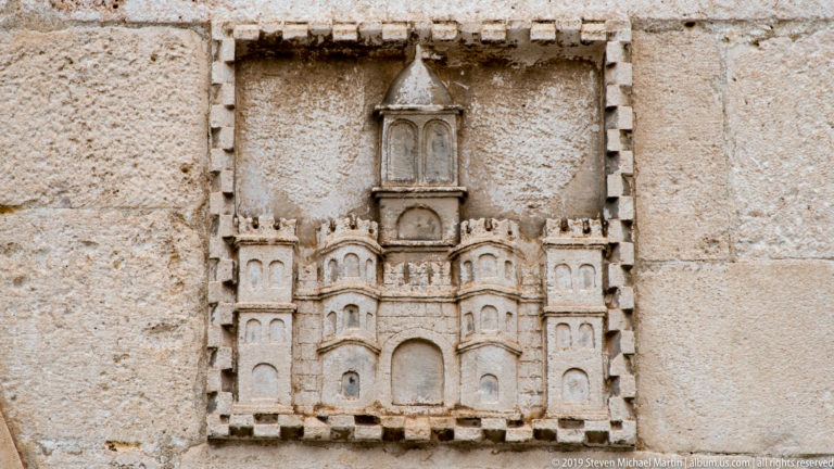 Small pale-stone relief of a symmetrical towered façade embedded in rough masonry.