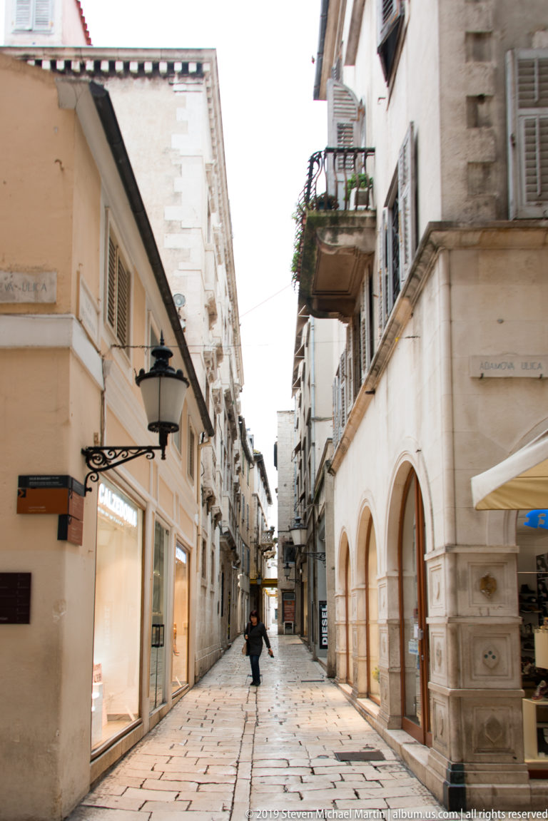 Narrow stone-paved pedestrian street lined with shops, shutters, balconies, and pale historic façades.