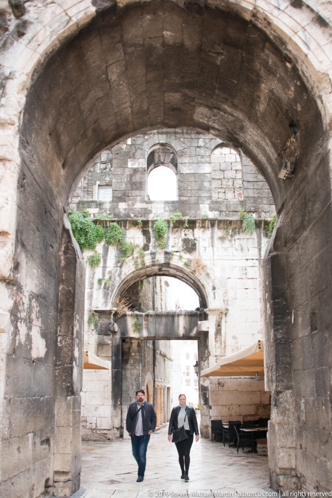 Two pedestrians walk beneath a massive stone arch leading through layered masonry passages.