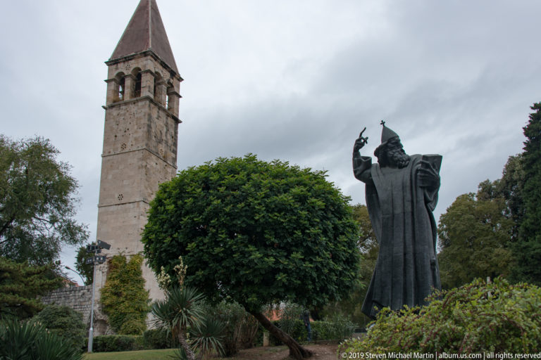 Dark bronze Gregory of Nin statue in a garden with a tall square stone bell tower behind.