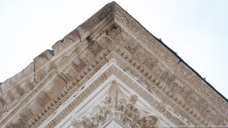 Upward view of a pale stone cornice densely carved with foliage, dentils, rosettes, and geometric bands.
