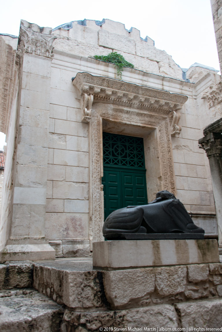 Reclining dark stone sculpture before an ornate pale-stone doorway with green doors in Split.