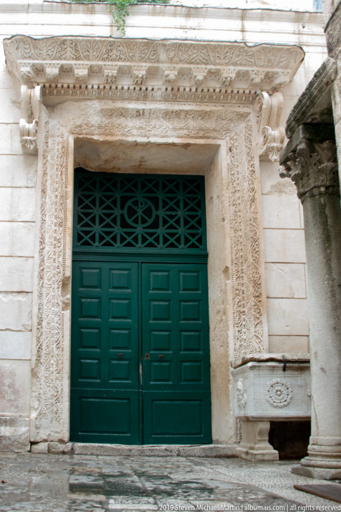 Ornately carved pale-stone portal surrounding green double doors with a patterned grille above.