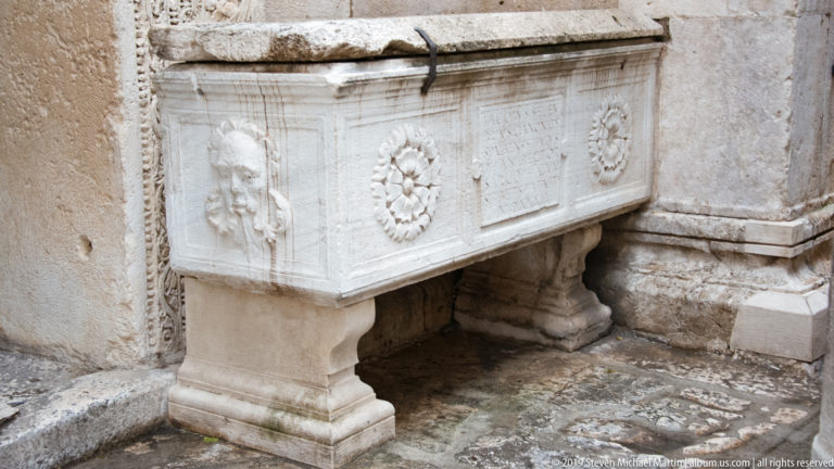 Carved pale-stone furnishing with relief faces, floral rosettes, inscription panel, pedestal supports, and weathered masonry behind it.