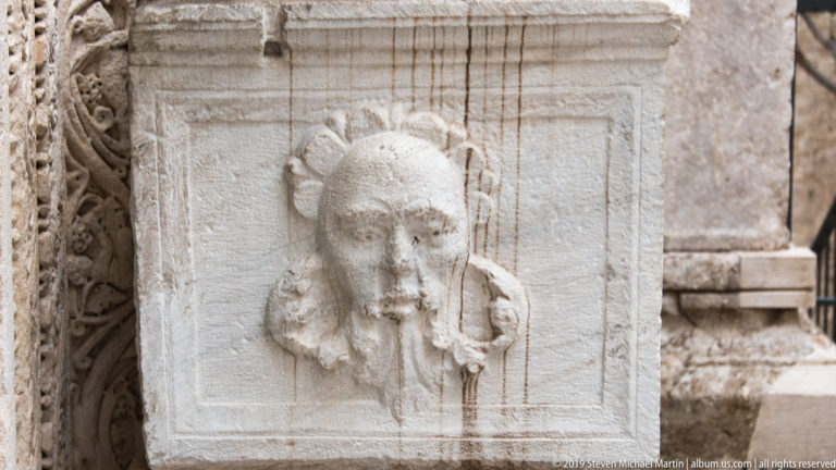 Close-up of a weathered carved human face projecting from a rectangular pale-stone panel with textured hair and worn features.