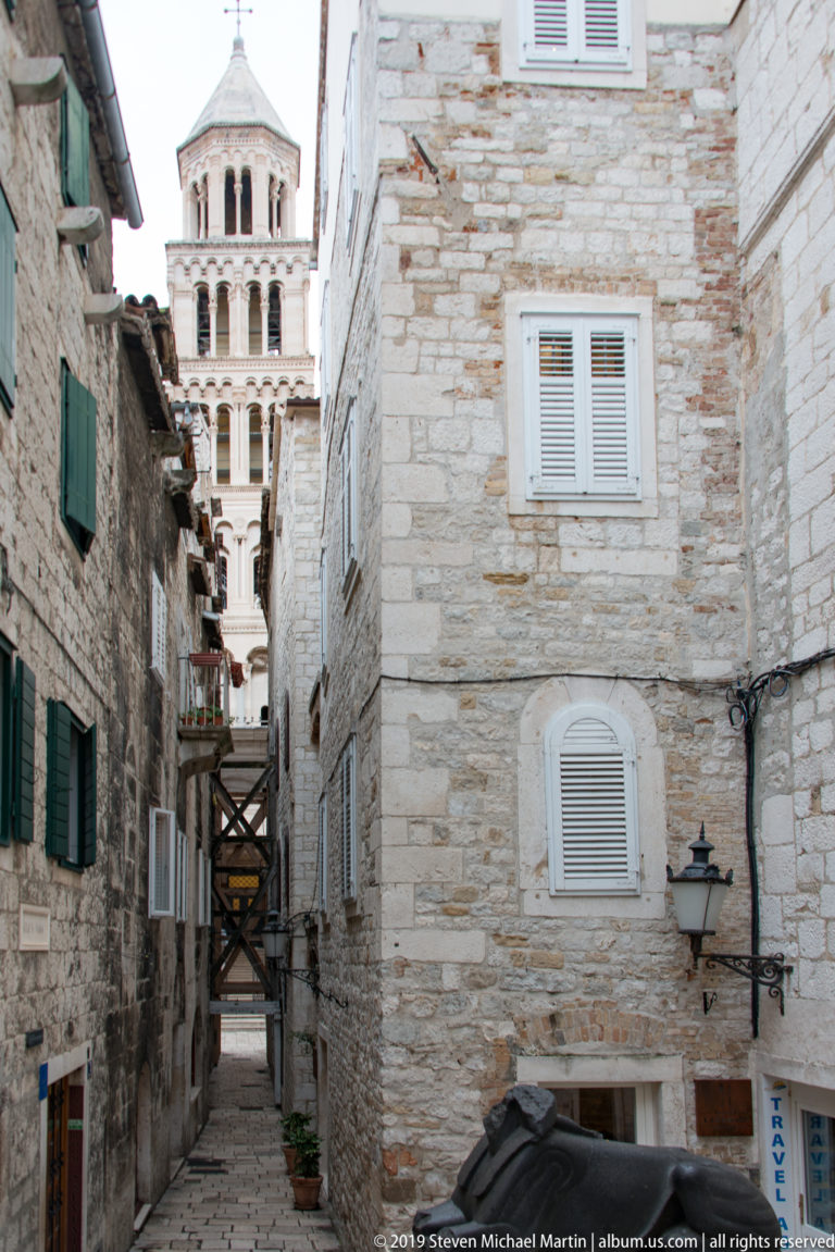 Narrow stone lane between shuttered buildings, with timber bracing and a partial cathedral bell tower visible at the far end.