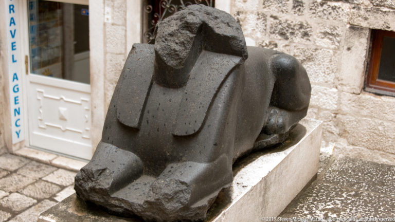 Dark recumbent stone animal sculpture with blocky head and forelegs on a pale rectangular plinth beside a stone building.