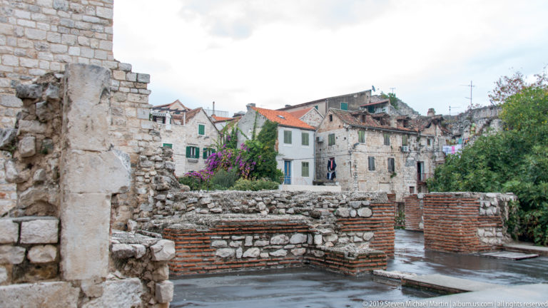 Low brick-and-stone archaeological walls in foreground with old houses, tiled roofs, greenery, and purple flowers behind.