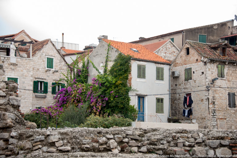 Old stone and stucco houses with green shutters and red roofs behind low ruins, with vivid purple bougainvillea in the center.