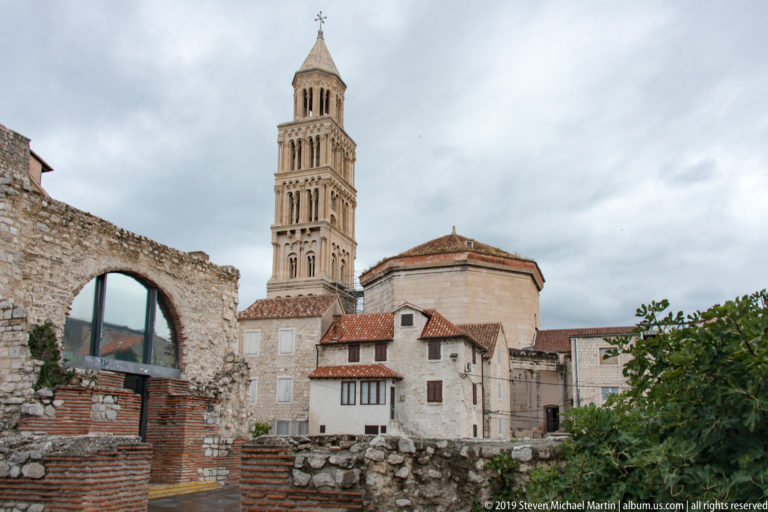 Cathedral bell tower and octagonal church seen beyond a broken stone arch and low brick-and-stone archaeological remains.