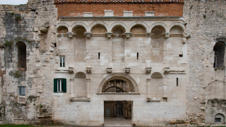 Pale symmetrical façade with central arched doorway, small windows and upper row of shallow arches beneath brickwork.
