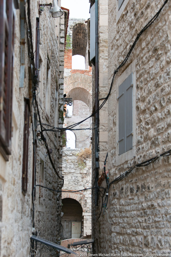 Narrow stone alley with rough masonry walls, gray shutters, overhead cables, brick repairs, and arches spanning the passage.