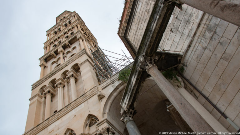Low-angle view of the Cathedral of Saint Domnius bell tower above stone arches and columns, with scaffolding alongside.