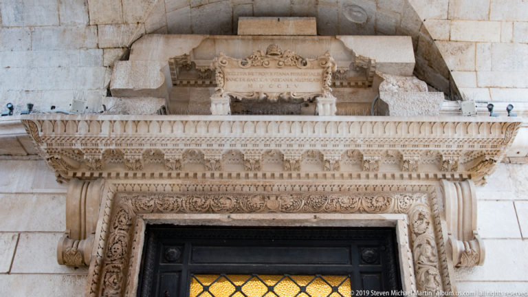 Ornate pale-stone doorway with carved foliate surround, projecting cornice, Latin inscription plaque, dark door, and amber glazing.