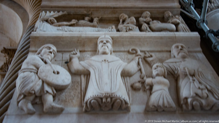 Low-angle close view of a carved stone relief with a central frontal figure, surrounding attendants, and smaller figures above.
