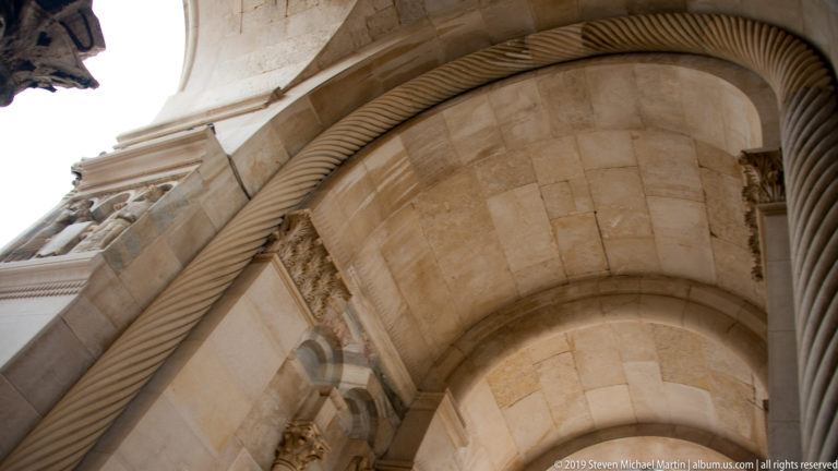Upward close view of a pale stone arch with thick twisted molding, carved capitals, and layered voussoirs.