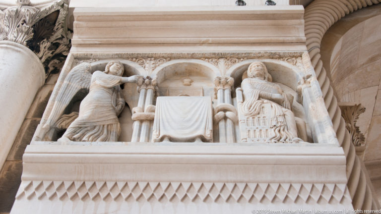 Carved stone relief with two large figures, central draped panel, miniature arches and columns, and diamond-pattern ornament below.
