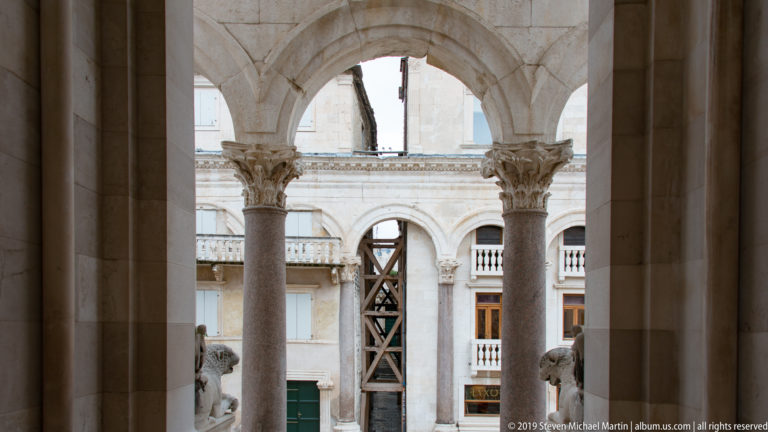 View through a large stone arch and two columns toward another arcade, façade, scaffolding, and partial lion sculptures below.