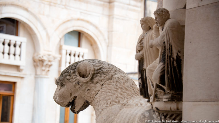 Carved stone lion in foreground with standing sculpted figures and cathedral architectural details behind it.