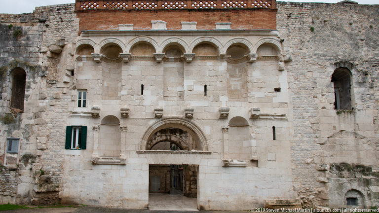 Pale historic stone façade with arched doorway, small windows and upper blind arches set within rough masonry.
