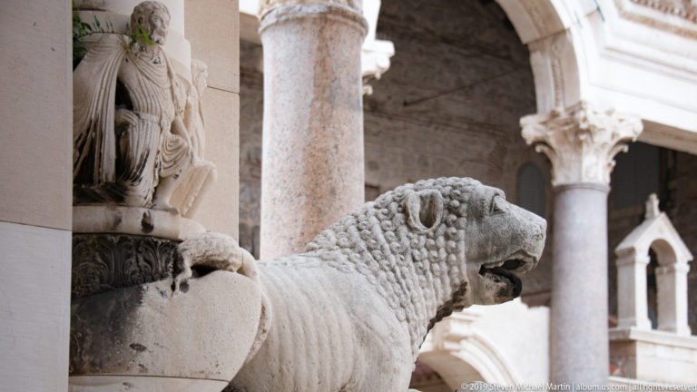 Close view of a carved stone lion beside a column, with curled mane, open mouth, polished shaft, and carved capital behind.