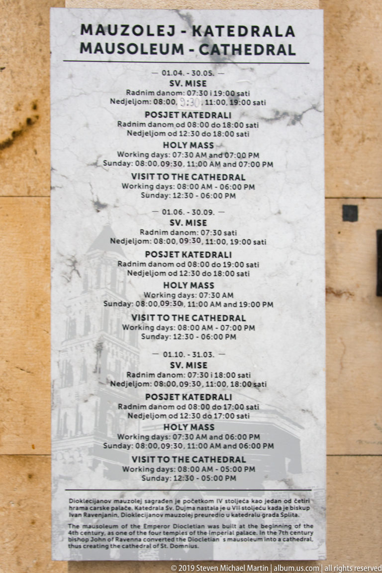 White multilingual cathedral notice listing Mass schedules and visitor hours in several languages on a stone wall.