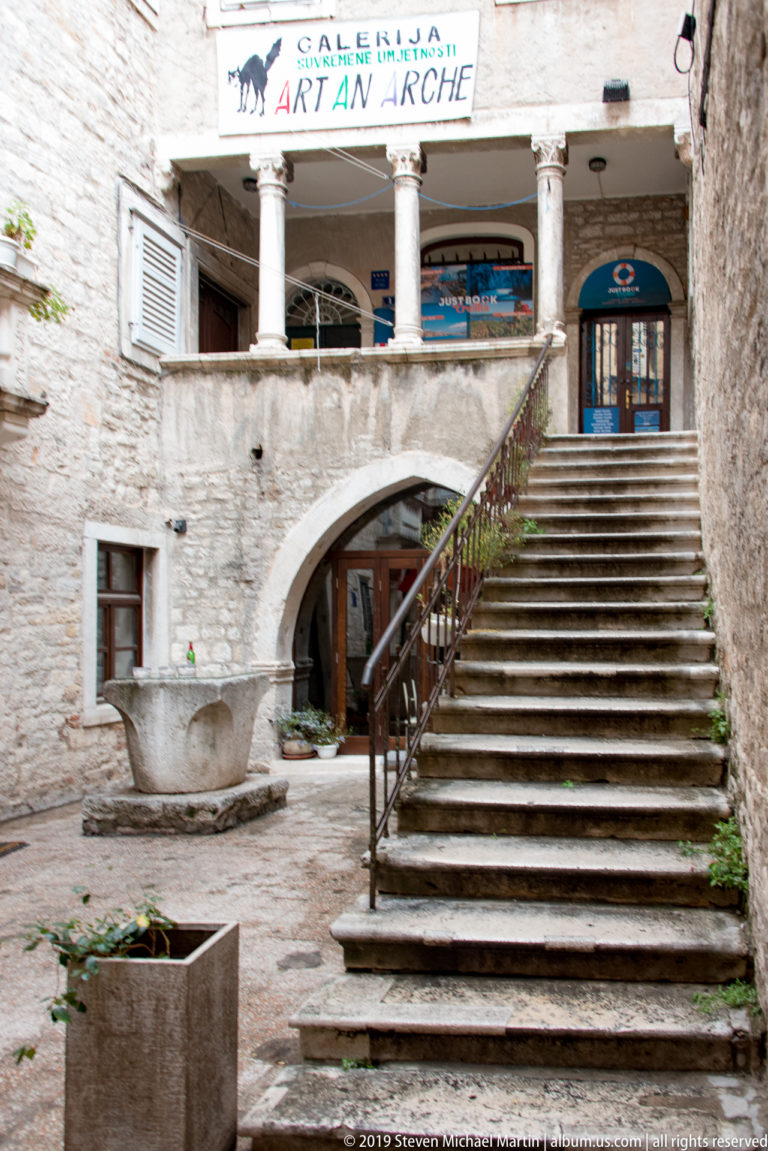 Wide stone staircase rising through a rough-masonry courtyard beneath an upper arcade, with gallery signage and modern doors.