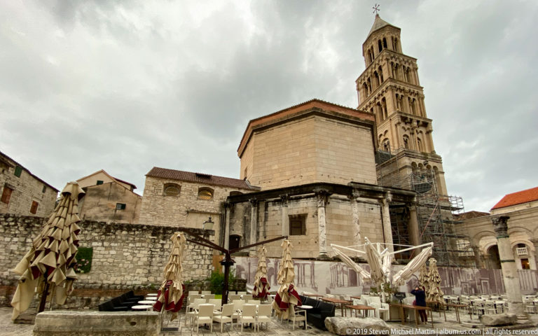 Octagonal cathedral structure and tall bell tower behind rough stone walls, café umbrellas, and archaeological masonry under an overcast sky.