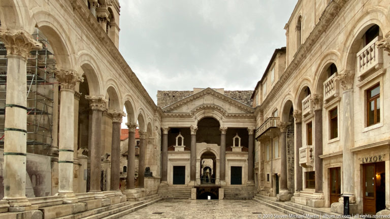Long stone-paved Peristyle court lined by columns and arcades, with a monumental arched façade centered at the far end.