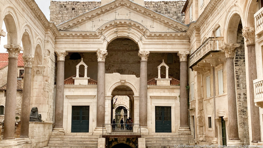 Symmetrical stone façade across the Peristyle with tall columns, central archway, side doors, balconies, and people on the steps.