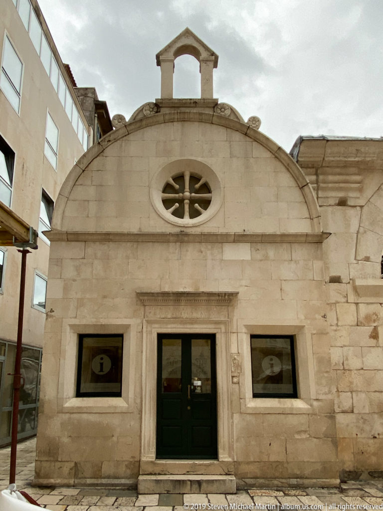 Small pale-stone gabled façade with central door, rectangular lintel, wheel-shaped rose window, and tiny bell opening above.
