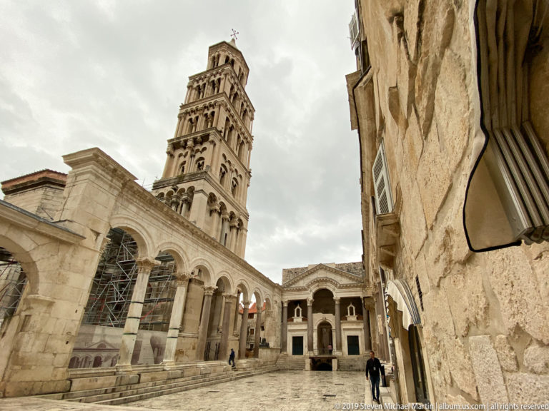 Cathedral bell tower rising above Peristyle arcades, with ancient columns at left and a weathered stone wall at right.
