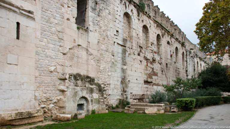 Long pale stone palace wall with repeated arches, rough masonry repairs, grass and trimmed shrubs.