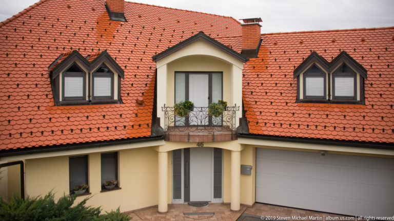 Cream detached house with broad red tile roof, three dormer groups, small balcony, arched entrance, and garage.