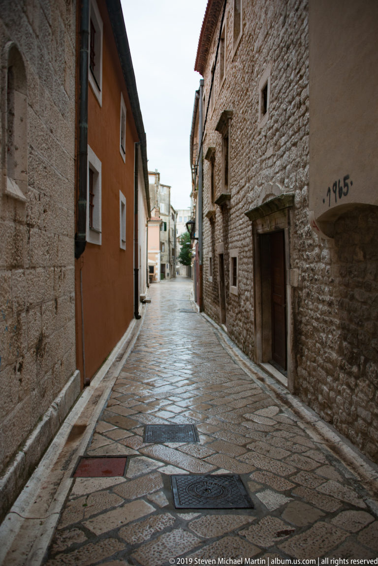 Narrow stone-paved lane between rough masonry and plastered houses with small windows and recessed doorways.