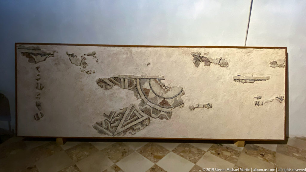 Fragmentary floor mosaic with dark geometric bands and curved motifs displayed against a pale background.