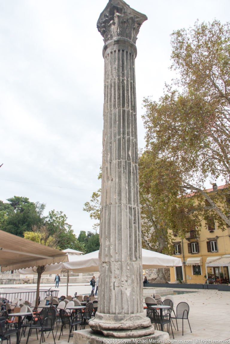 Tall weathered fluted stone column with classical capital standing among café tables and awnings in a public square.