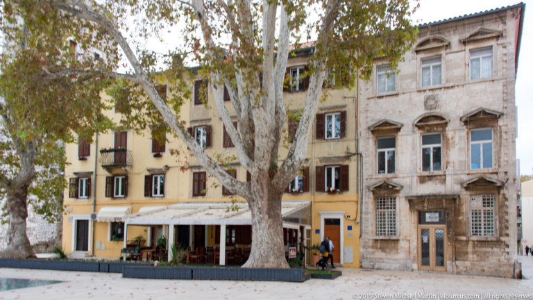 Weathered pale-stone townhouse with tall windows and carved lintels partly screened by a large street tree beside a yellow building.