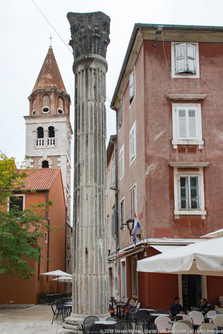 Fluted ancient stone column with carved capital in front of a red building and historic bell tower.
