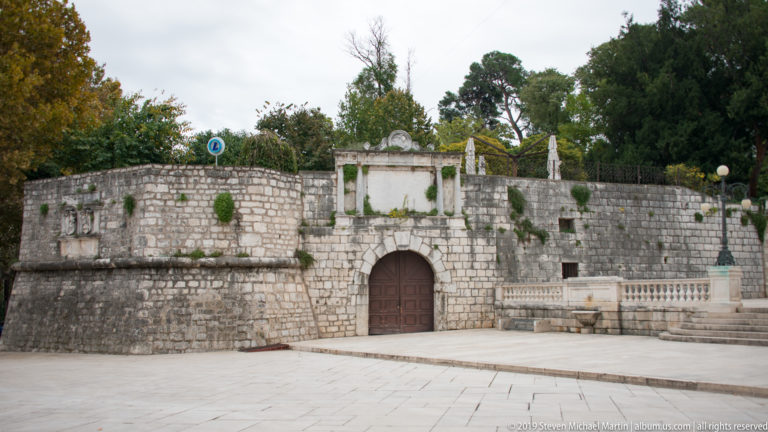 Round-arched wooden gate in a curved stone fortification wall with balustrades, trees, and landscaping above.