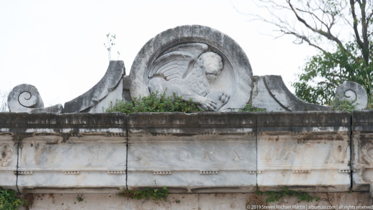 Long weathered stone monument with central circular figural relief and large curling volutes at both ends.