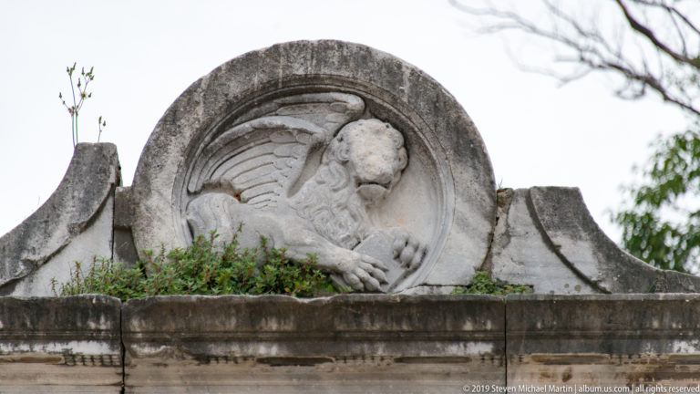 Weathered circular stone relief with a reclining winged or allegorical figure above an overgrown ledge.
