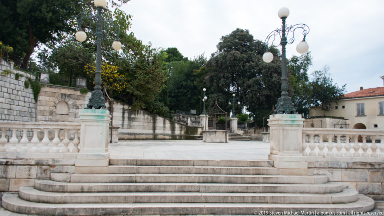 Broad stone steps with balustrades and ornate lamp posts leading toward a tree-filled terrace above Five Wells Square.