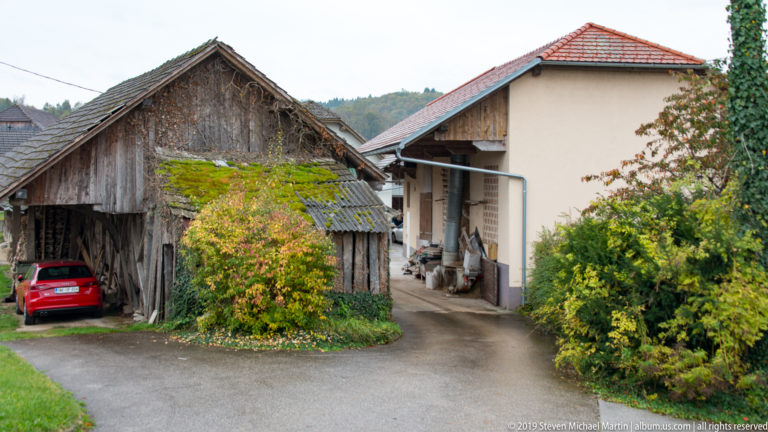 Weathered wooden farm outbuildings beside a stuccoed house, with vines, shrubs, a narrow drive, and red car.