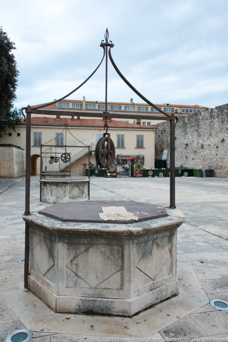 Polygonal stone wellhead with decorative iron frame and pulley wheel in Zadar’s Five Wells Square.